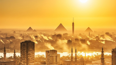 صورة بانورامية لمدينة القاهرة تظهر فيها الأهرامات أو برج القاهرة في الأفق، مع شمس كبيرة ومتوهجة في السماء. أهم شيء في الصورة هو وجود تموجات حرارية (Heat Haze) واضحة تتصاعد من المباني أو الأسفلت، مما يعطي إحساسًا بصريًا قويًا بالحرارة الشديدة. الألوان تميل إلى الأصفر والبرتقالي.